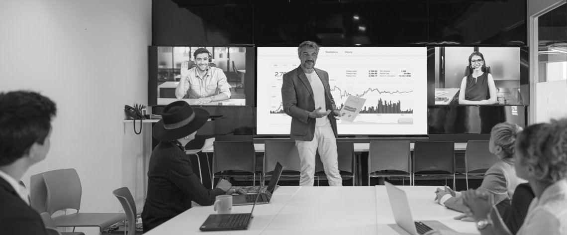 A man presents in a glass meeting room with conference room sound. Several audience members sit at the meeting table, other participants can be seen on screens attending the meeting.